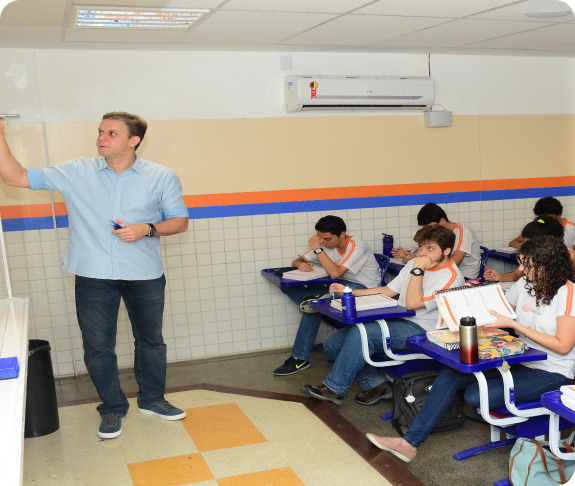 Professor apontando para a lousa explicando o conteúdo, com alunos sentados em carteiras ao fundo prestando atenção