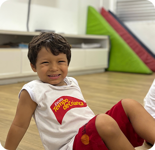 Menino sorrindo sentado no chão vestindo camiseta Tempo de Criança
