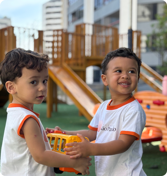 Dois meninos sorridentes brincando com um caminhão de brinquedo amarelo em um parquinho ao ar livre, vestindo camisetas do Colégio São Paulo