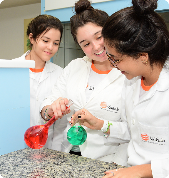 Três estudantes de jaleco branco fazendo experiência de química com frascos coloridos, vermelho e verde, em laboratório escolar