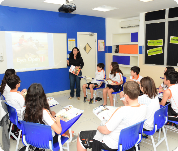 Professora em pé dando aula para turma sentada em cadeiras azuis, com tela de projeção ao fundo em sala de aula com paredes azul e branco